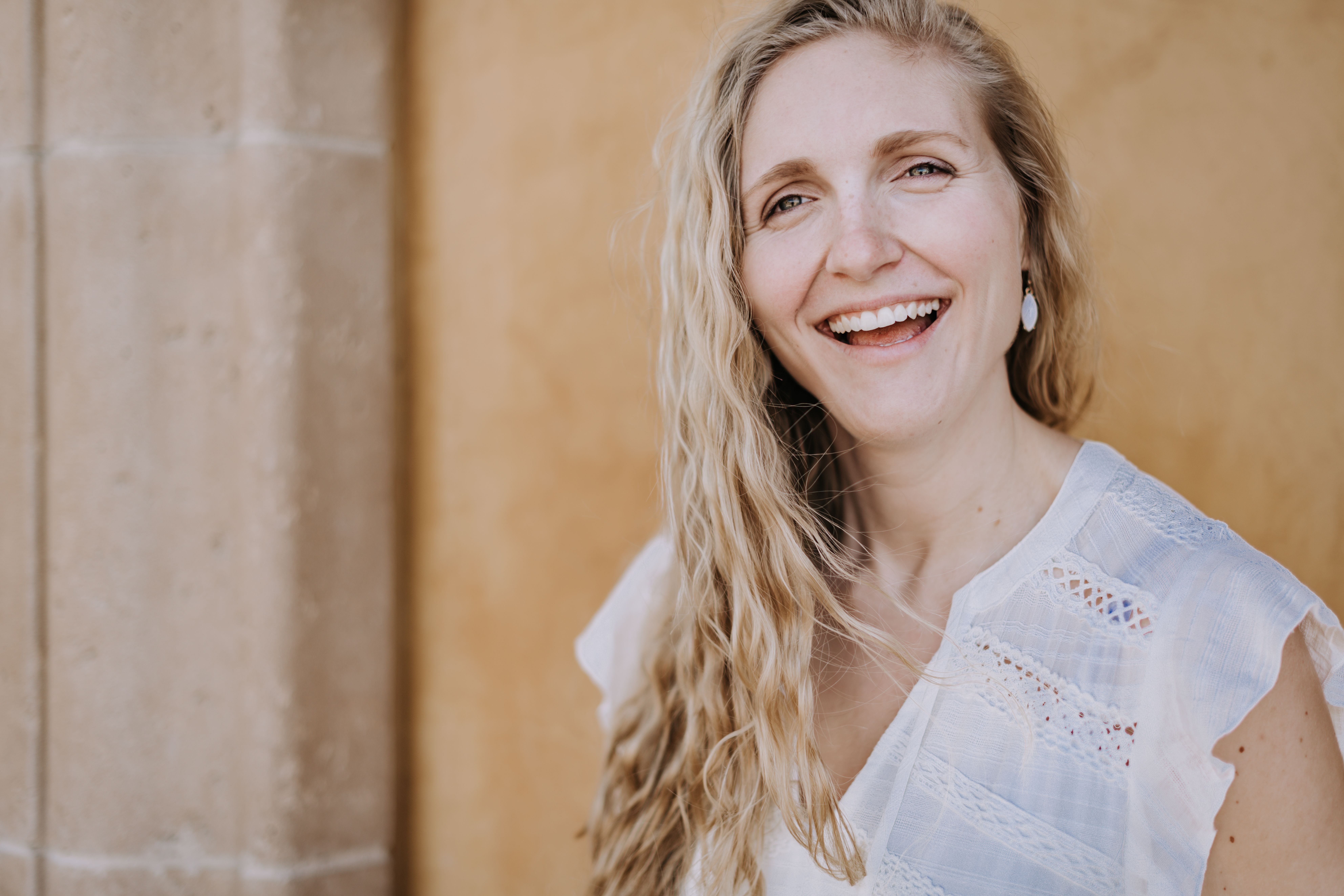 A cheerful woman with long hair and a bright smile, standing against a textured wall.
