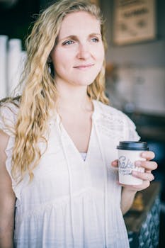 Woman holding a coffee cup indoors, smiling softly in natural light.