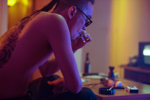 Shirtless man with tattoos smoking indoors, surrounded by moody lighting and urban decor.