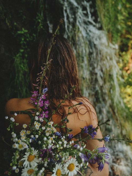 Free Woman standing by a waterfall holding vibrant wildflowers, creating a serene blend of nature and beauty. Stock Photo