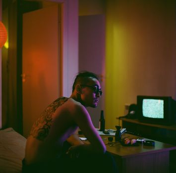 A shirtless man with sunglasses and tattoos sits in a dimly lit room with moody lighting.