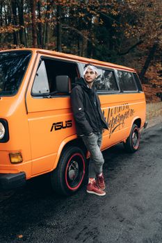 Casually dressed man leaning on an orange van, parked by a forest road during fall.