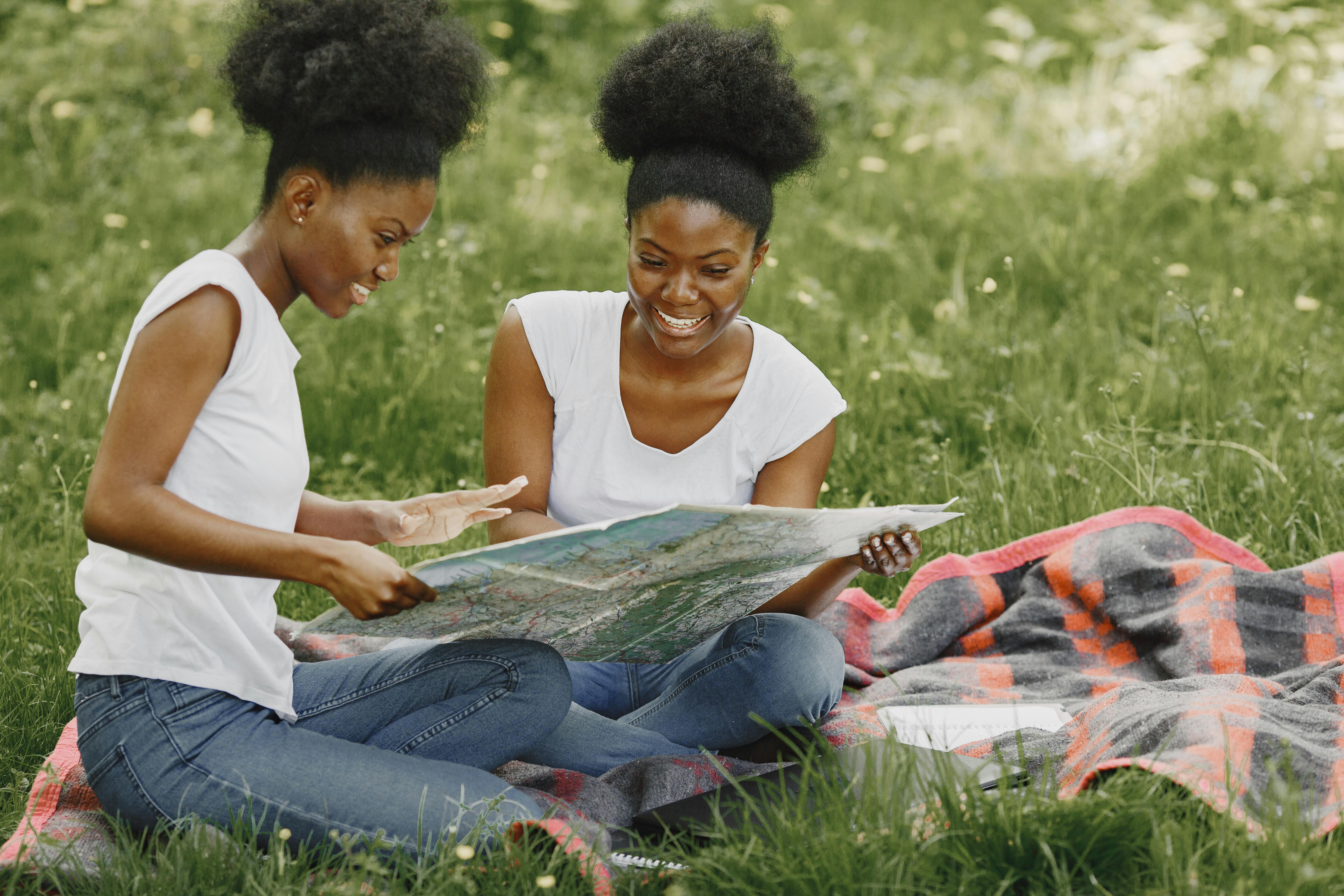 Free Two women sitting on a picnic blanket in a grassy area, looking at a map and smiling. Stock Photo