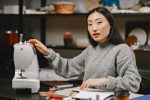 Free Asian woman dressmaker in a cozy atelier working with a sewing machine, representing small business and creativity. Stock Photo