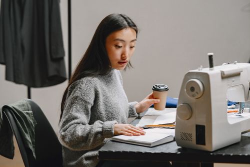 Free Asian woman designing clothes in a modern workspace, holding coffee, and using a sewing machine. Stock Photo