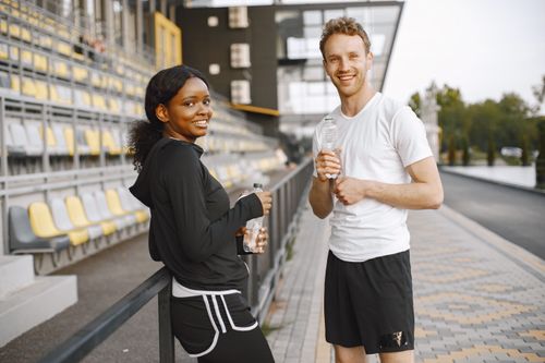 Free A cheerful couple in sports attire enjoying a break outdoors holding water bottles, perfect for lifestyle themes. Stock Photo