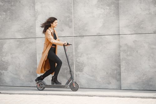 Free A woman in a brown coat rides an electric scooter against a modern concrete wall. Stock Photo