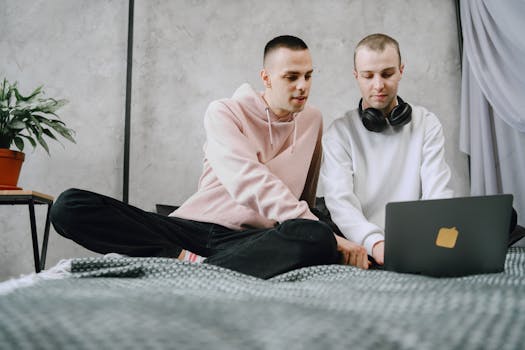 Two adults working on a laptop while sitting on a bed indoors, wearing casual clothes.