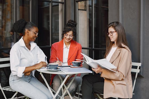 Three women in diverse business attire having a meeting outdoors with coffee and documents.
