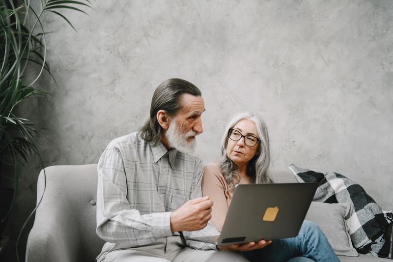 Elderly couple engaged in discussion while using a laptop at home.