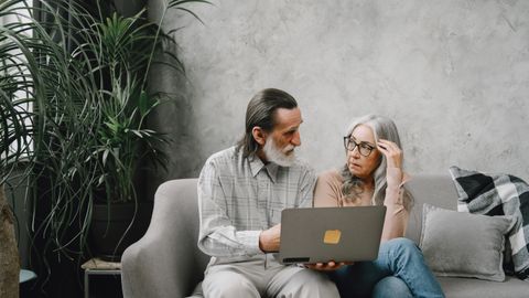 Elderly couple discussing while using laptop at home on sofa, surrounded by plants.