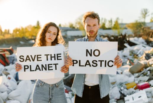 Two young adults hold signs against plastic pollution in a landfill, urging environmental action.