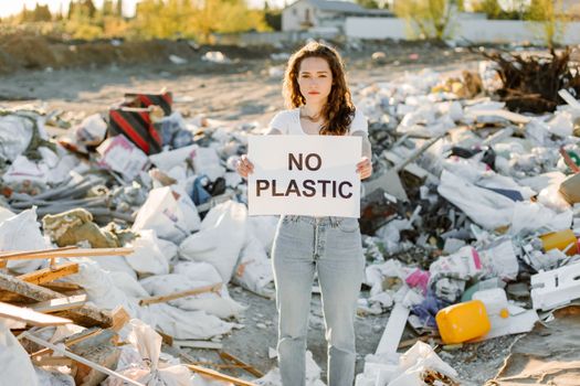 A young woman holds a sign saying 'No Plastic' in a junkyard to support environmental awareness.