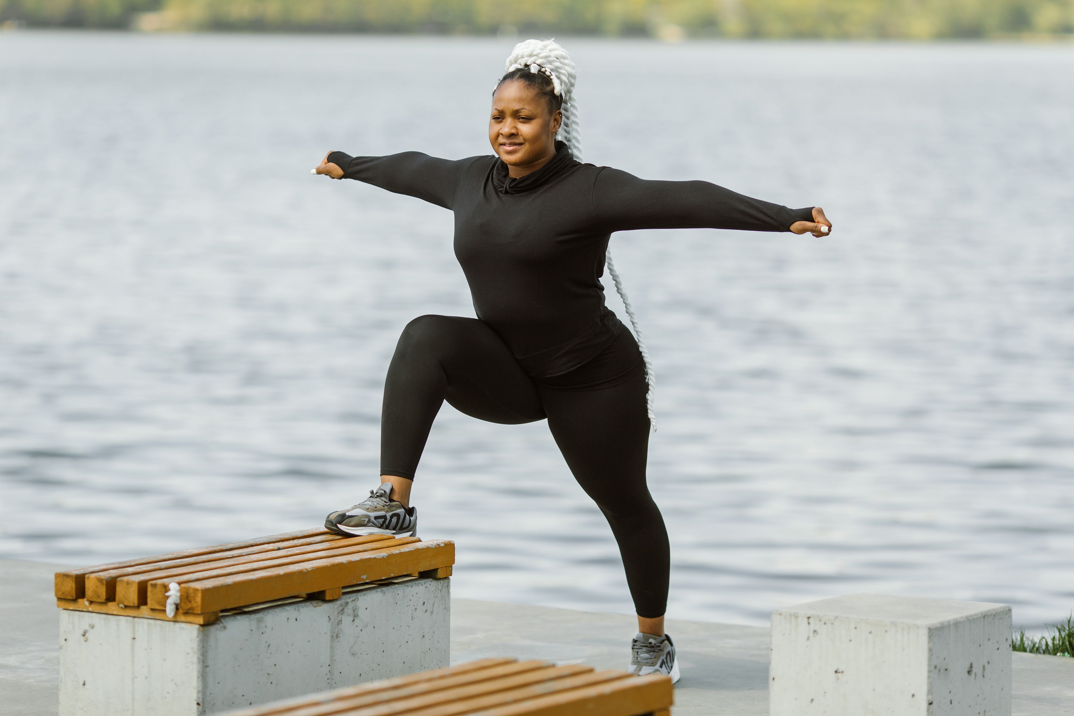 A plus size black woman in activewear stretching outdoors by a serene lake.