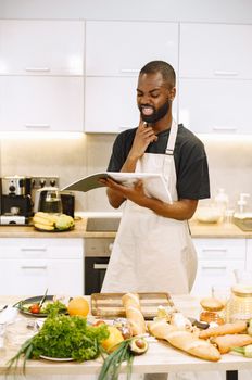 A man in an apron reads a recipe book in a bright kitchen with various ingredients.
