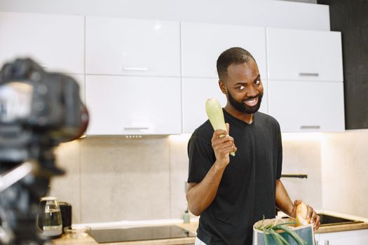 Smiling black man prepares meal in kitchen for vlog, holding zucchini.
