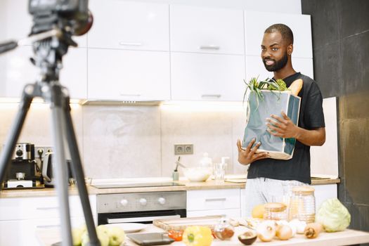 A man recording a cooking vlog in a modern kitchen holding a grocery bag.