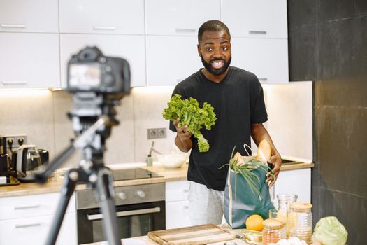 A man vlogging in a kitchen with fresh vegetables and recording equipment, showcasing a cooking segment.