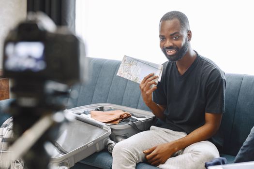 Smiling young man recording a travel vlog at home with camera and open suitcase.