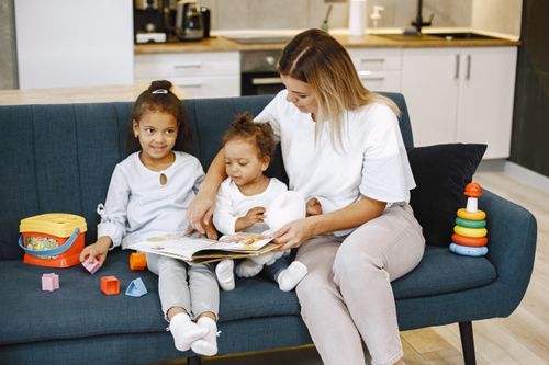 Free A mother reads to her two daughters on a couch, surrounded by colorful toys, indoors. Stock Photo
