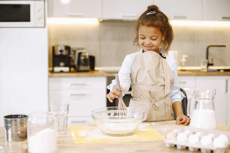 Girl With Apron Standing Behind The Kitchen Counter Mixing Flour For Baking