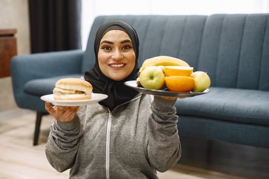 A smiling woman wearing a hijab holds a burger and a plate of fruits in a cozy indoor setting.