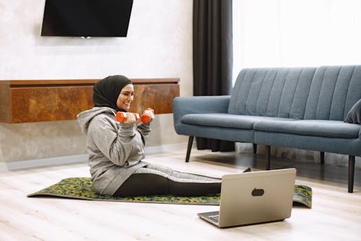 A woman in hijab exercises with dumbbells at home, sitting on a mat, engaging in a virtual workout session.