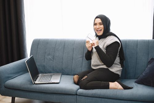 Free Smiling woman in hijab using a laptop on sofa, shopping online. Indoor lifestyle scene. Stock Photo