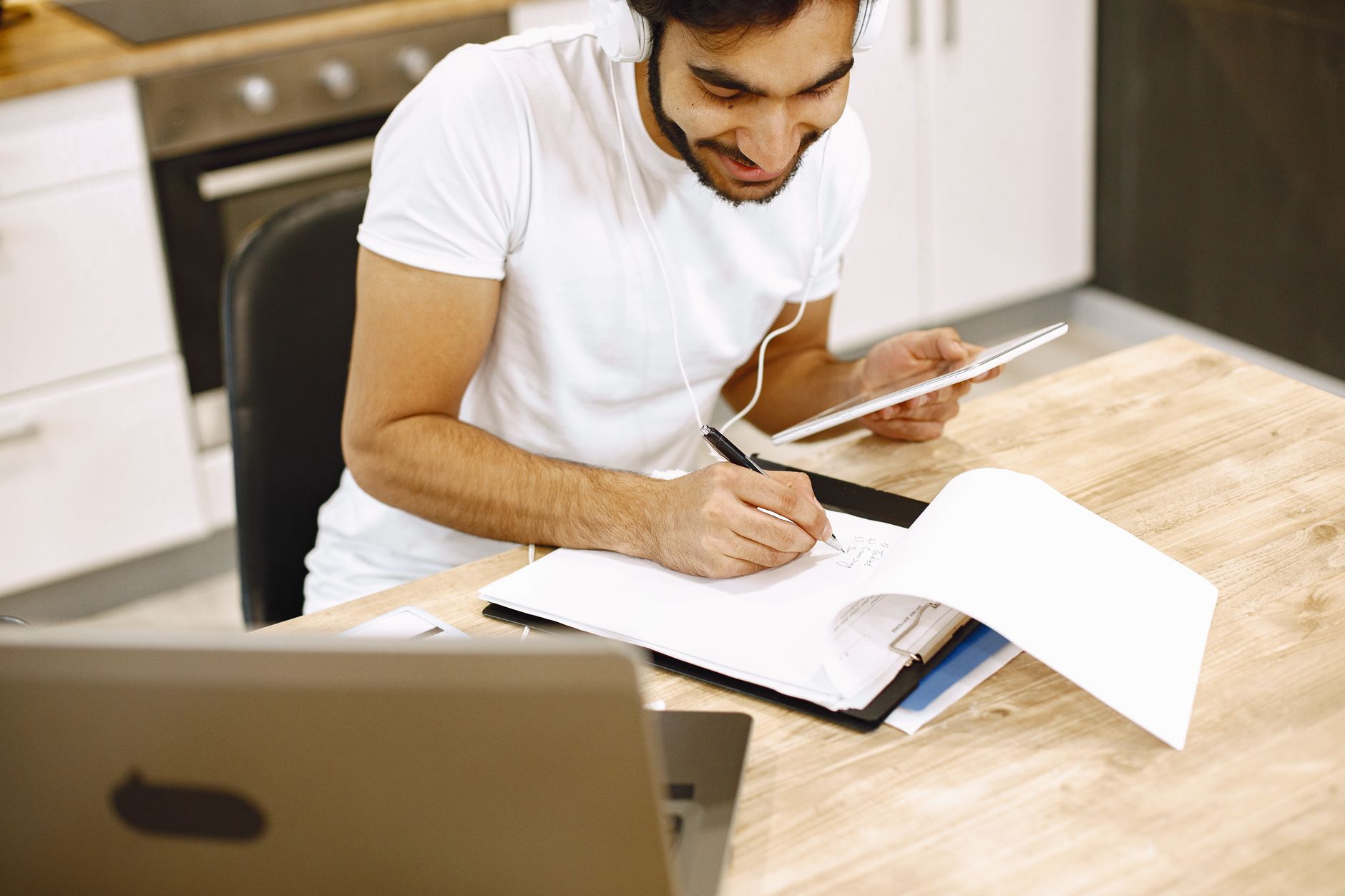 Adult man takes notes while using tablet and laptop in a modern kitchen setting.