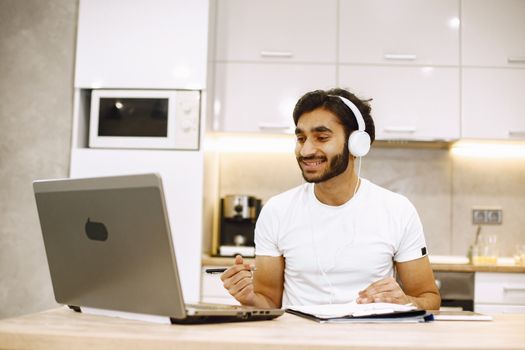 Smiling young man wearing headphones working on a laptop in a modern home kitchen.