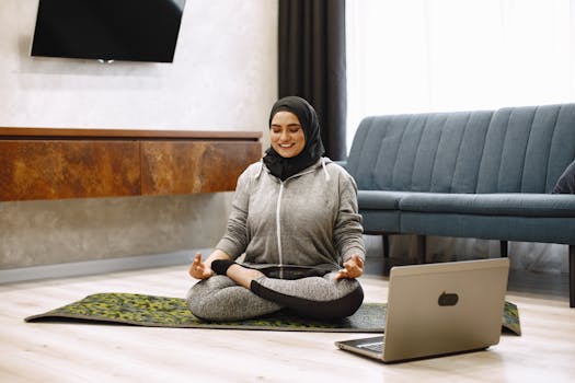 A young woman practicing yoga and meditation indoors, sitting on a yoga mat with a laptop nearby.