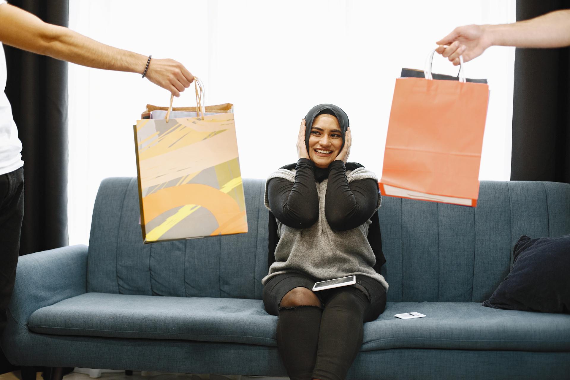 A cheerful woman on a couch receiving gift bags while smiling indoors.