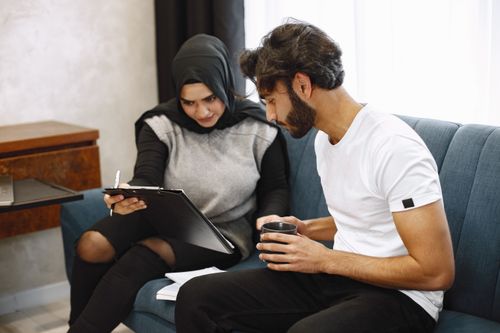Free Middle Eastern man and woman on sofa, collaborating with clipboard and coffee. Stock Photo