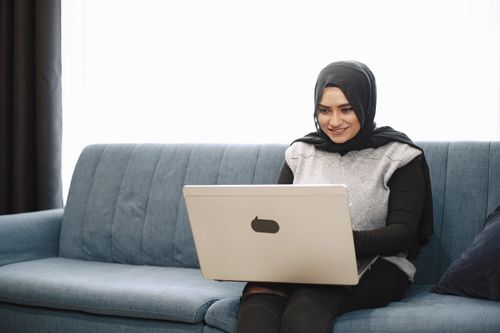 Free Smiling woman in hijab sitting on a sofa using a laptop, enjoying leisure time indoors. Stock Photo
