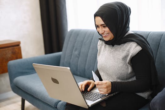 A woman in a hijab smiles, working on a laptop from a comfortable sofa.