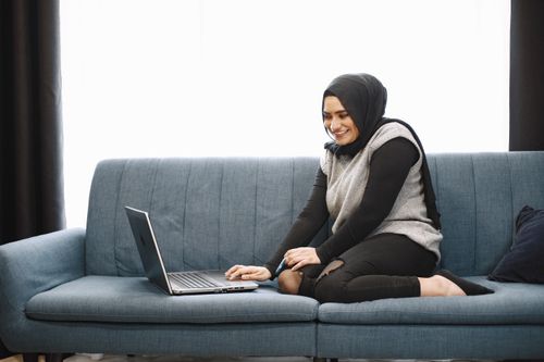 Free A smiling Middle Eastern woman in a hijab using a laptop on a blue sofa indoors. Stock Photo