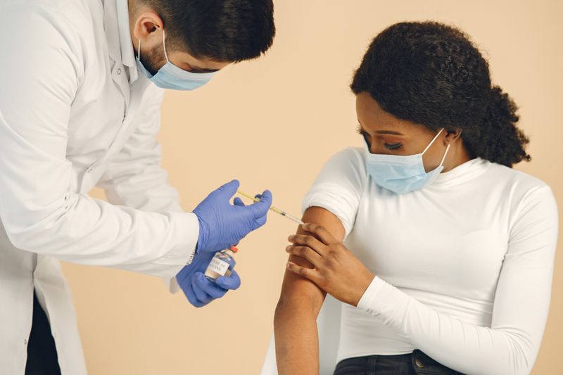 A Black woman receives a seasonal flu vaccine in her upper arm from a masked clinician wearing gloves, in a clinical setting.