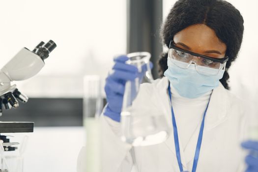 Woman scientist in lab coat and mask conducting experiment with microscope and beaker.