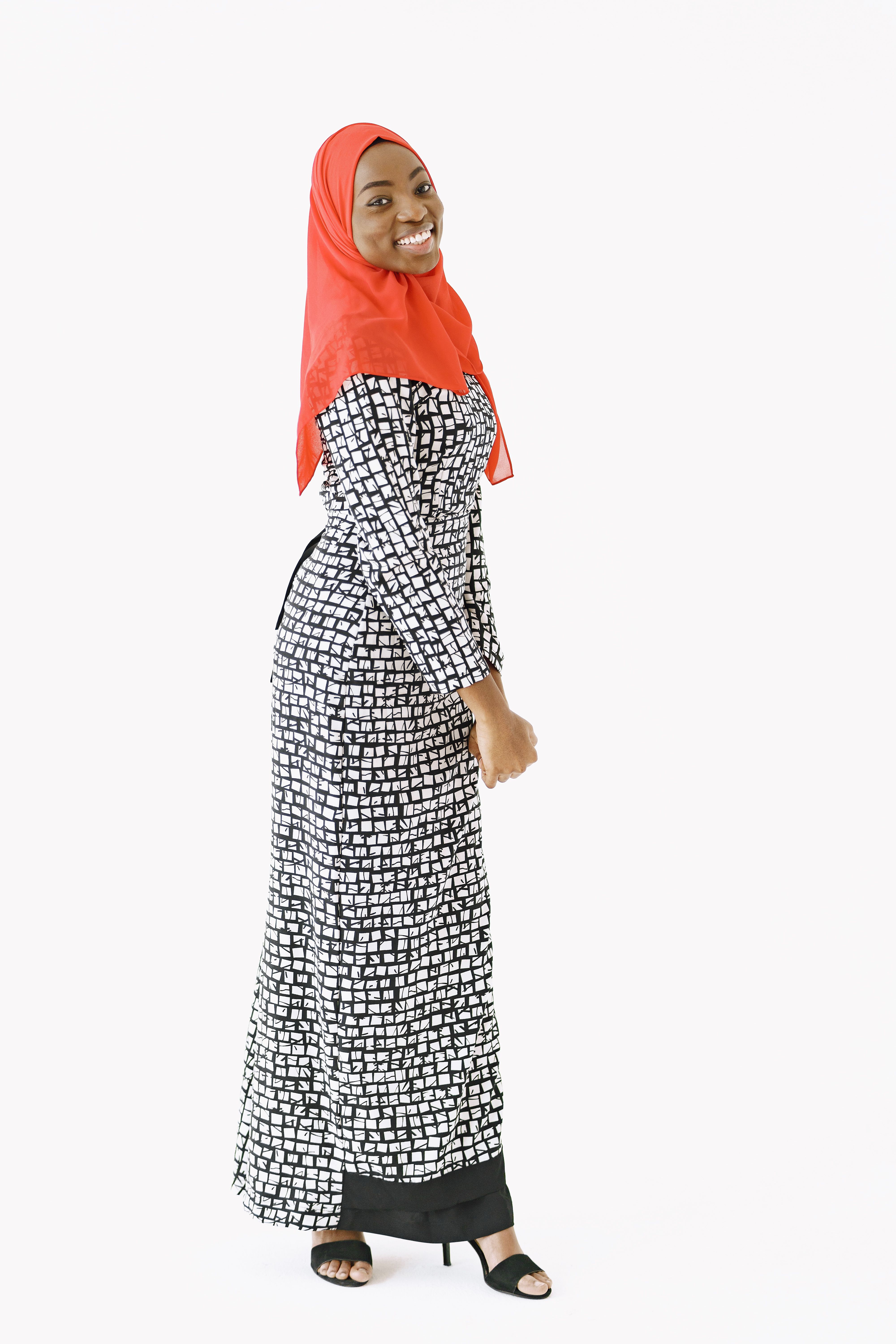 Smiling woman in black and white dress with red hijab, standing on a white background.