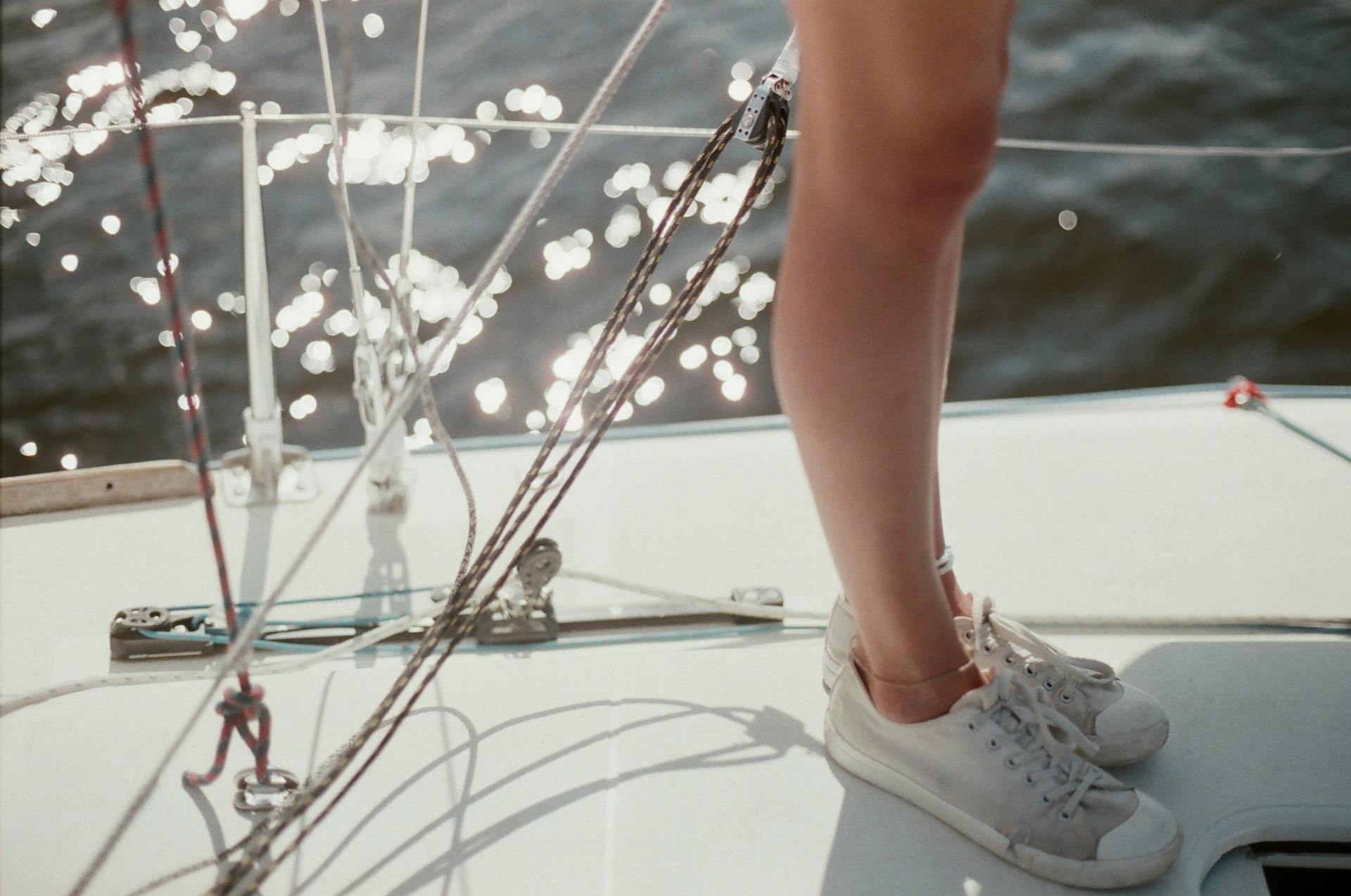 A woman's legs in sneakers are seen standing on the deck of a sailboat with sparkling water in the background.