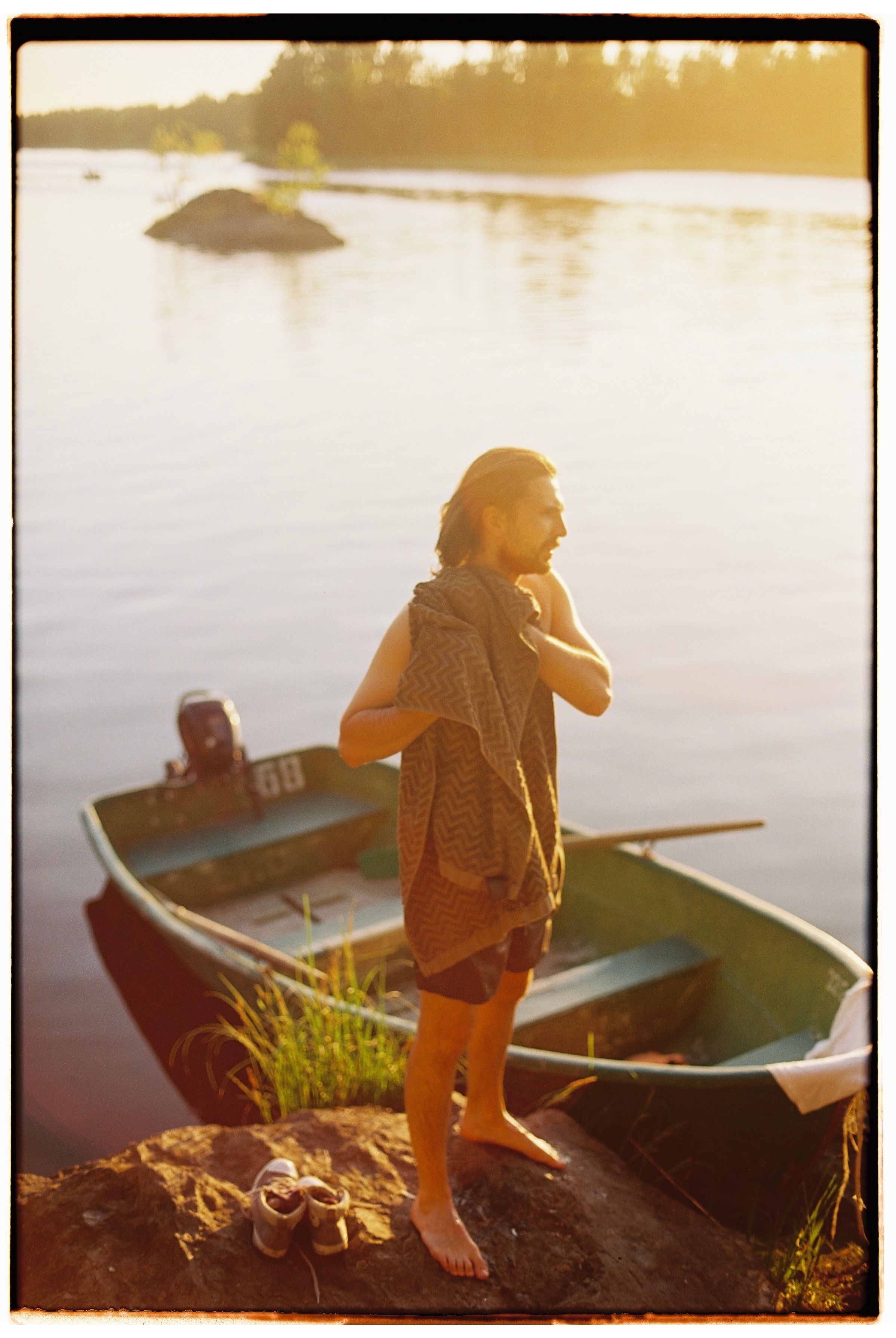 A man stands riverside with a boat in the background, enjoying a sunset moment.