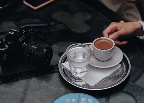 Free A Canon camera beside a ceramic coffee cup and glass of water on a metallic tray. Stock Photo