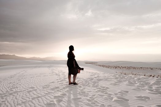 A woman stands in the serene desert landscape at sunset, surrounded by sand dunes.