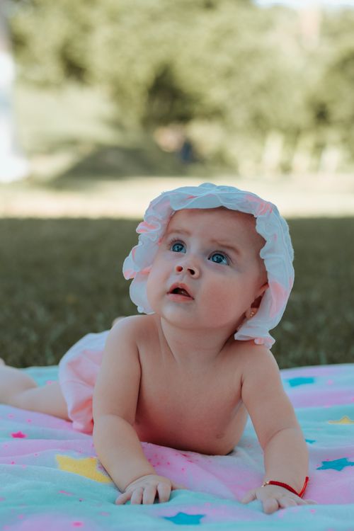 Free Cute baby wearing a sun hat, gazing upward while lying on a colorful blanket in a sunny outdoor setting. Stock Photo