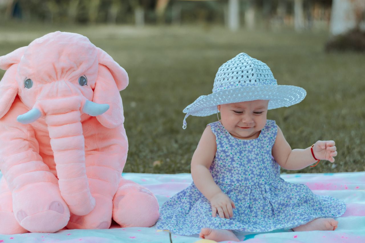 adorable baby sitting on a blanket outdoors wearing a hat next to a pl