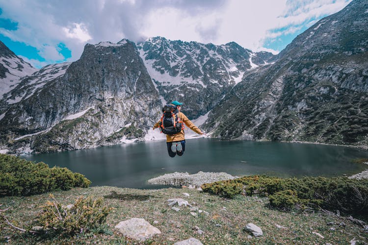 A Person Wearing Backpack Jumping Near The Lake