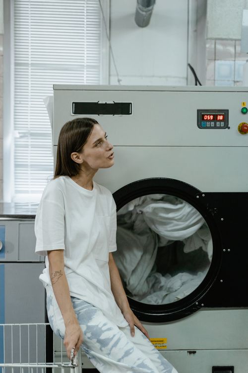 Free Woman sitting by washing machine in a bright, modern laundromat. Casual and relaxed atmosphere. Stock Photo
