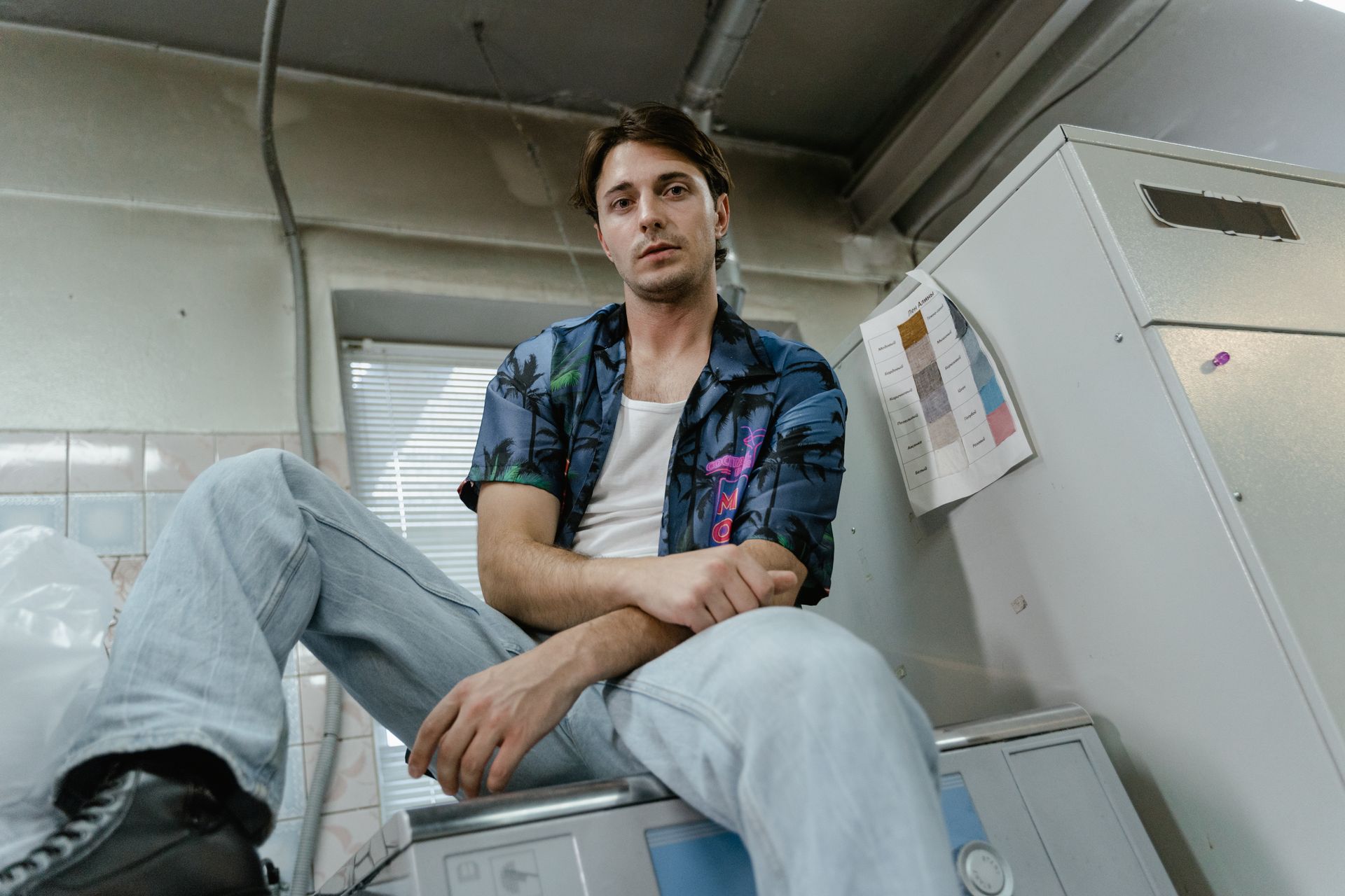 A man in a printed shirt sits on a washing machine in an indoor laundry room setting.