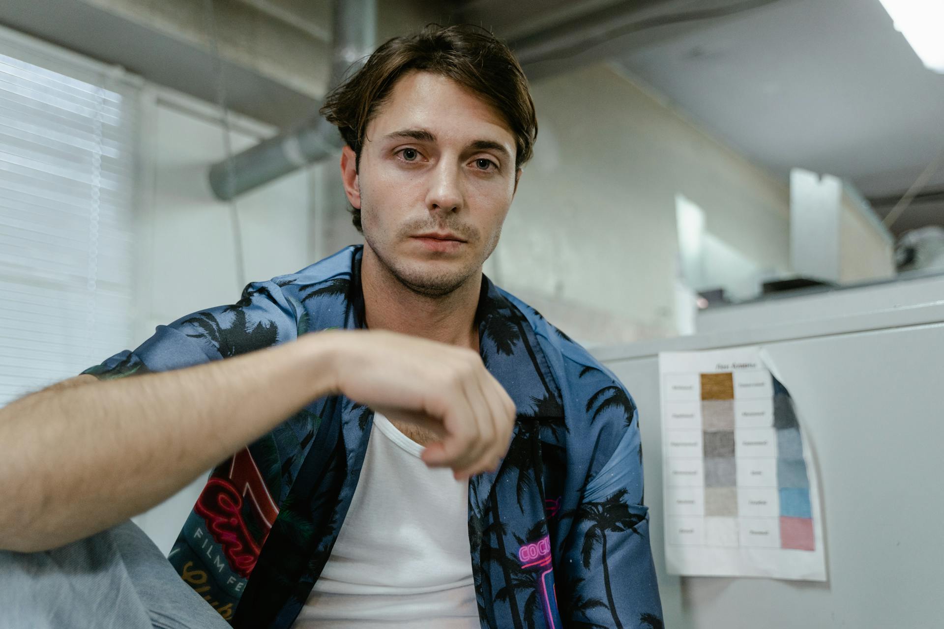 Young man wearing an aloha shirt, sitting indoors with a casual expression.