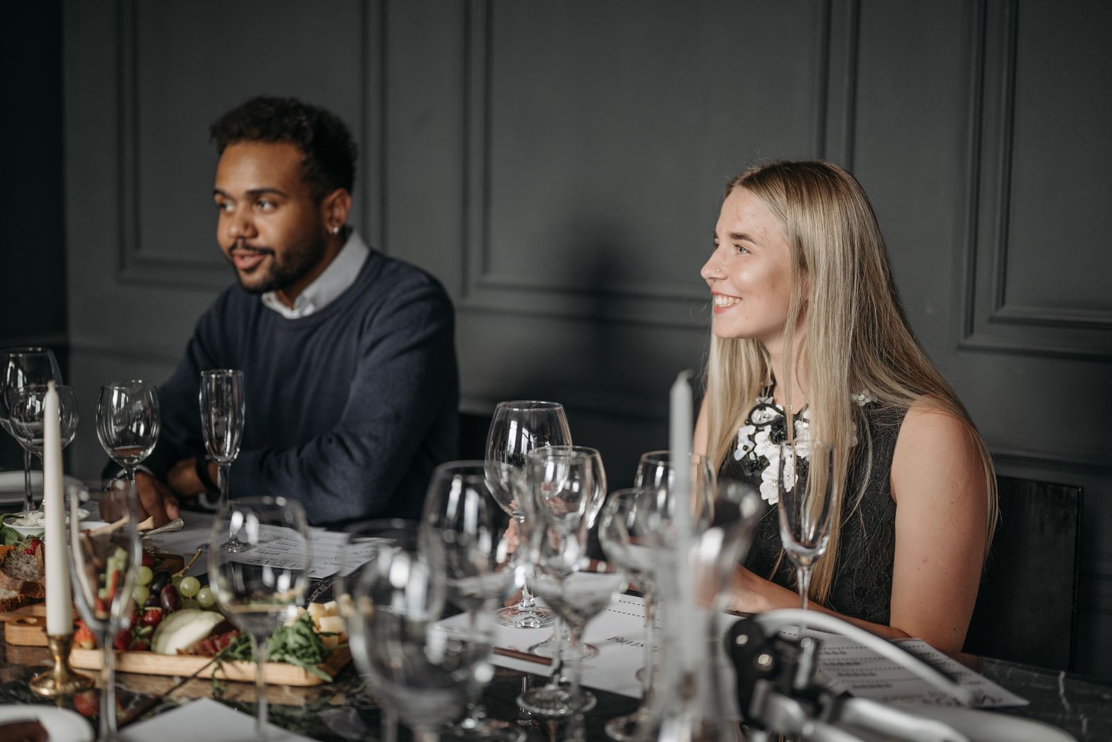 Two guests in profile, mid-conversation across a candlelit table in a dark panelled room
